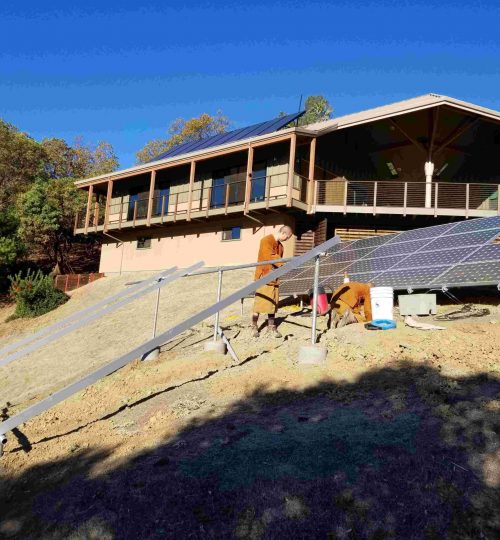 Two monks working to construct two large solar panels on a hillside in front of a larger building